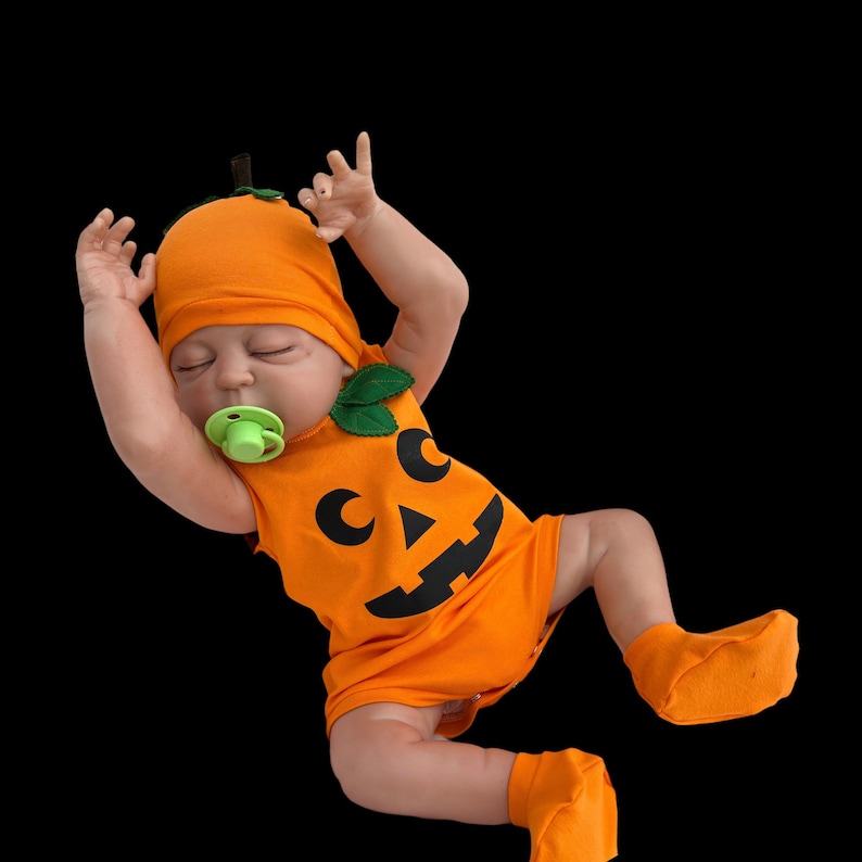 May include: A baby doll wearing an orange pumpkin costume with a green leaf and black facial features. The doll is wearing a pacifier.