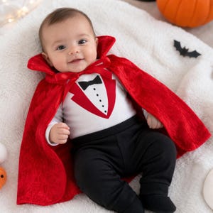 May include: A baby wearing a white long-sleeve bodysuit with a printed black bow tie and red tuxedo lapel design, paired with solid black pants. A vibrant red velvet cape with a high collar and a matching red ribbon tie is worn over the outfit.