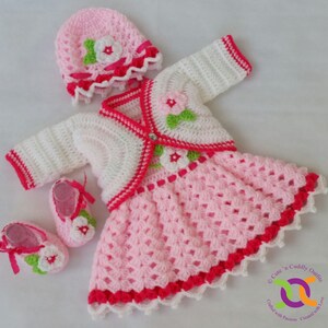Op de afbeelding: Een gehaakte baby-outfitset in roze en wit met een bijpassende muts en schoentjes. De outfit bestaat uit een jurkje met een wit vest, roze bloemen en een rode bies. De muts heeft een roze bloem en een wit lint. De schoentjes hebben een roze bloem en een wit lint.
