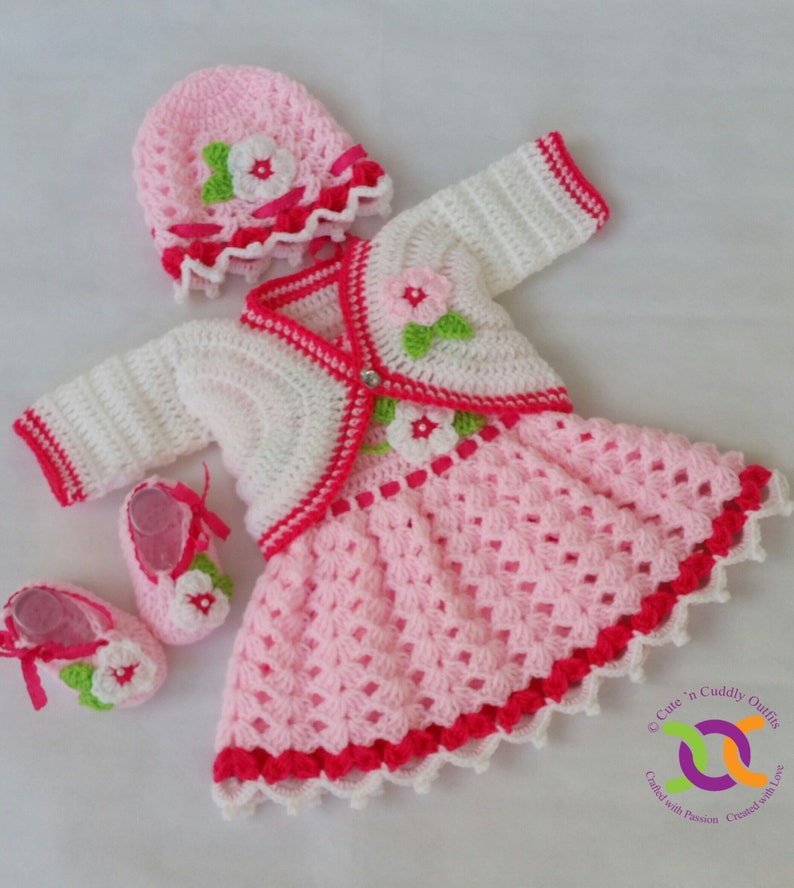 Op de afbeelding: Een gehaakte baby-outfitset in roze en wit met een bijpassende muts en schoentjes. De outfit bestaat uit een jurkje met een wit vest, roze bloemen en een rode bies. De muts heeft een roze bloem en een wit lint. De schoentjes hebben een roze bloem en een wit lint.