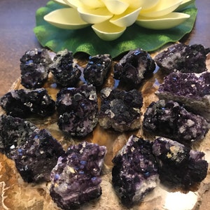 May include: A collection of raw fluorite crystals in shades of purple and black, arranged on a brown, marbled surface. The crystals have a rough, natural texture and are illuminated by soft lighting. A white and yellow flower is in the background.