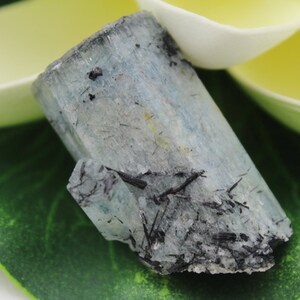 Puede incluir: Un cristal de aguamarina azul claro con inclusiones de turmalina negra. El cristal es rugoso y tiene un aspecto natural sin pulir.