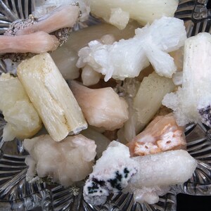 May include: A collection of white and pink selenite crystals in a clear glass bowl. The crystals are various shapes and sizes, some are long and thin, others are short and stubby.