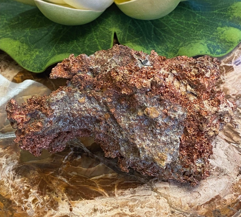 Puede incluir: Una roca grande, rugosa, de color cobre con un brillo met&aacute;lico. La roca tiene una variedad de texturas y formas, con algunas &aacute;reas que parecen m&aacute;s lisas y otras m&aacute;s irregulares. La roca est&aacute; descansando sobre una superficie marr&oacute;n y texturizada.