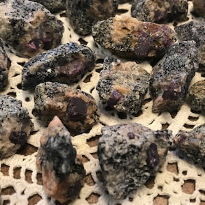 May include: A collection of small, rough, dark gray and brown rocks with embedded red crystals. The rocks are arranged on a white crocheted surface.