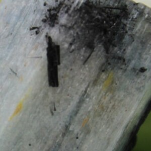 Puede incluir: Primer plano de una muestra mineral de color azul claro y gris con inclusiones negras. El mineral tiene una superficie estriada y una textura rugosa.
