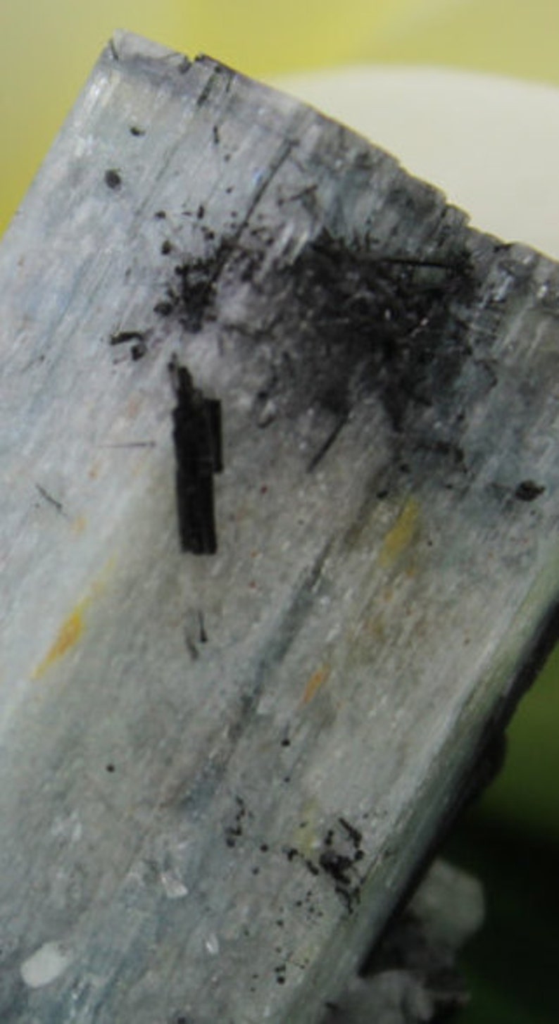 Puede incluir: Primer plano de una muestra mineral de color azul claro y gris con inclusiones negras. El mineral tiene una superficie estriada y una textura rugosa.
