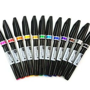 Puede incluir: Un conjunto de 12 marcadores negros de punta fina con tinta de colores. Los marcadores están dispuestos en forma de abanico con las puntas hacia afuera. Los colores de la tinta son rosa, naranja, amarillo, verde, azul, morado, marrón, plata y oro.