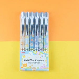 Puede incluir: Un paquete de seis bolígrafos de gel EnerGel Kawaii. Los bolígrafos tienen clips plateados y están decorados con bonitos diseños de animales en colores pastel. El embalaje es blanco con el texto "EnerGel Kawaii" y una ilustración de oso. El fondo es naranja y amarillo.