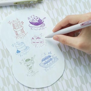 May include: A white paper sheet featuring hand-drawn illustrations of various desserts in different ink colors, including a chocolate bar, sundae, cake, pancakes, parfait, and dango, each labeled with Japanese text and color names.