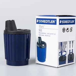 May include: A blue Staedtler Mars 502 pencil sharpener with a black top and a clear plastic window. The sharpener is in its original packaging, which is white with blue and black text. The packaging shows a close-up of the sharpener and instructions on how to use it.