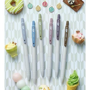 May include: Six Pilot Juice gel pens with light gray barrels and colored clips in blue, light blue, green, mauve, purple, and brown. The pens feature the text Juice and the number 5 on the clips. Small decorative charms and miniature food items are arranged around the pens.