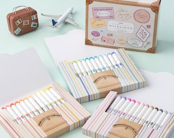 May include: Three sets of colorful water-based markers in striped boxes. The boxes are decorated with a travel theme and the text "Tombow  Voyage! Limited Edition." The markers are labeled "Tombow  Mildliner" and "Twin Tip".
