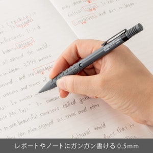 May include: A gray mechanical pencil with the text "SMASH 05 Pentel G1005 JAPAN" is held over a notebook. The pencil is being used to write on the lined paper. The text at the bottom says "レポートやノートにガンガン書ける 0.5mm".
