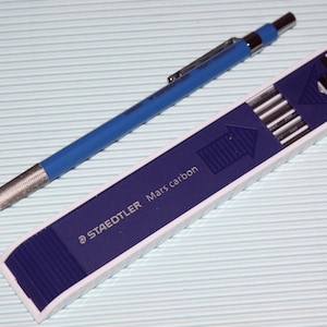 Puede incluir: Un lápiz mecánico azul con un agarre de metal plateado y un paquete de 12 minas de lápiz mecánico Staedtler Mars carbon en una caja de plástico azul y blanco. Las minas tienen un diámetro de 2 mm y están etiquetadas como "HB".