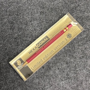 Peut inclure: Un crayon mécanique rouge et doré dans un emballage en plastique transparent. Le crayon est étiqueté "HEXAGONAL Mechanical pencil 0.5" et "Limited edition version-3". L'emballage porte le logo Staedtler.