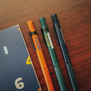 Op de afbeelding: Drie mechanische potloden met verschillende kleuren: oranje, groen en zwart. De potloden zijn gelabeld met "SMASH 05" en "Pentel Q1005 JAPAN".