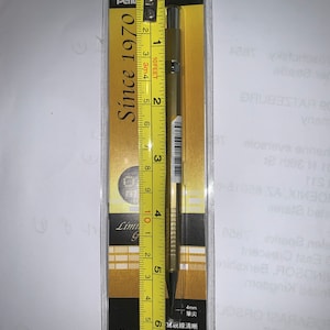 Könnte beinhalten: Ein goldener mechanischer Bleistift mit einem schwarzen Clip und einer silbernen Spitze. Der Bleistift ist in einem durchsichtigen Plastiketui mit goldenem Hintergrund verpackt, auf dem der Text "Since 1970" und "Limited Edition" aufgedruckt ist. Der Bleistift ist 14 cm lang.