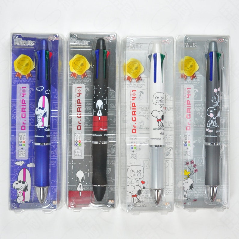 May include: Four multi-colored Dr. Grip 4+1 mechanical pencils with Peanuts characters. The pencils are blue, black, silver, and gray. Each pencil has a different Peanuts character on the barrel. The text "Dr. Grip 4+1" is printed on each pencil.