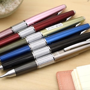 May include: A collection of six retractable ballpoint pens arranged in a row. Each pen features a metallic barrel in various colors including pink, red, olive green, blue, black, and charcoal, paired with a textured silver grip and clip.