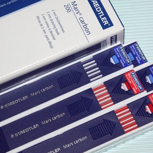 May include: A pack of Staedtler Mars Lumochrom drawing leads in blue and red packaging. The leads are 2 mm in diameter and are labeled "Mars Lumochrom 12 x 2.0 mm".
