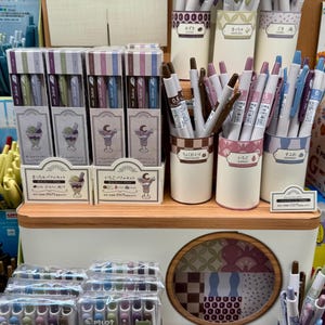 May include: A retail display of Pilot Juice gel pens in various colors, featuring boxed sets with parfait illustrations and individual pens in cylindrical holders labeled with Japanese text and color names.