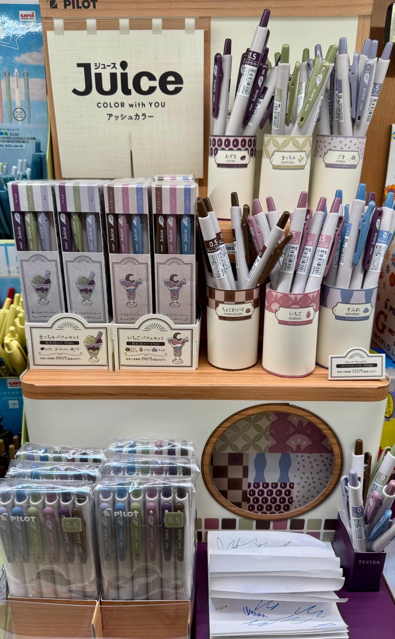 May include: A retail display of Pilot Juice gel pens in various colors, featuring boxed sets with parfait illustrations and individual pens in cylindrical holders labeled with Japanese text and color names.