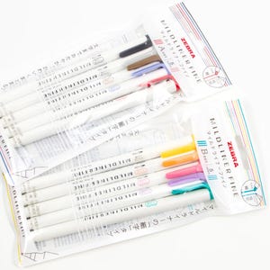 Pode incluir: Duas embalagens de marcadores Zebra Mildliner Fine. Cada embalagem contém um conjunto de canetas em várias cores, incluindo preto, castanho, azul, vermelho, amarelo, laranja, rosa, roxo e turquesa. A embalagem apresenta texto japonês e o logótipo Zebra.