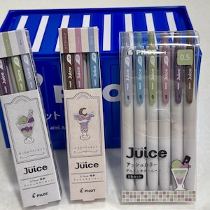 May include: A set of three Pilot Juice 0.5mm gel pen packages in ash colors. Two packages are vertical boxes with dessert illustrations, and one is a clear plastic case containing six pens with colored clips and white barrels, labeled Juice.