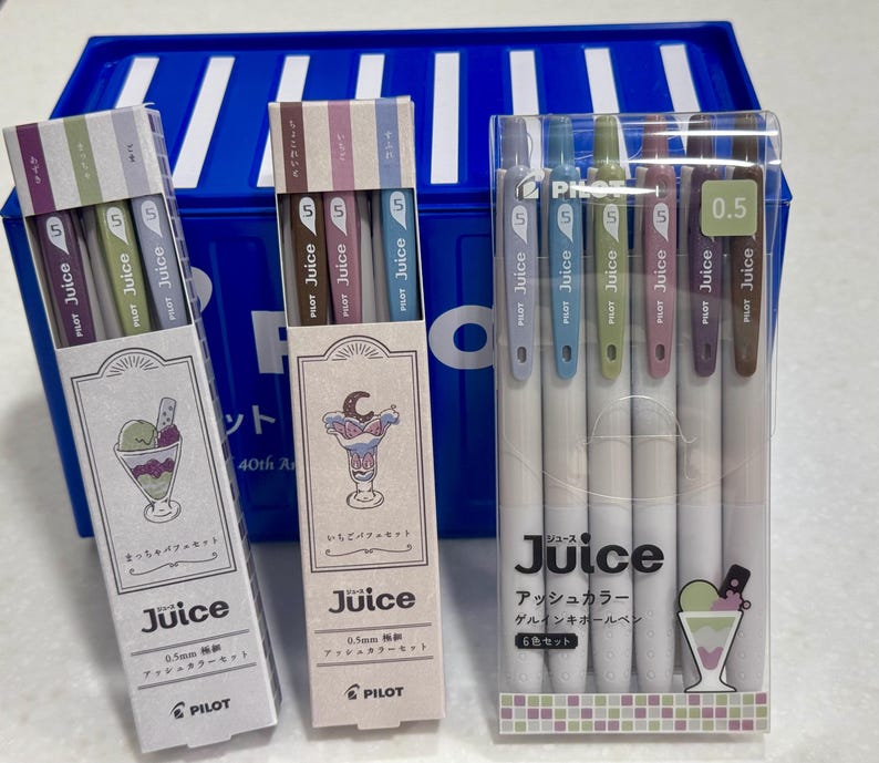 May include: A set of three Pilot Juice 0.5mm gel pen packages in ash colors. Two packages are vertical boxes with dessert illustrations, and one is a clear plastic case containing six pens with colored clips and white barrels, labeled Juice.