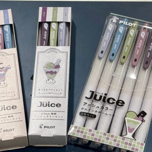 May include: Three sets of Pilot Juice 0.5mm gel ink pens in ash colors. Two sets are in vertical cardboard boxes with dessert illustrations, and one set is in a clear plastic case containing six pens with colored clips and white barrels.