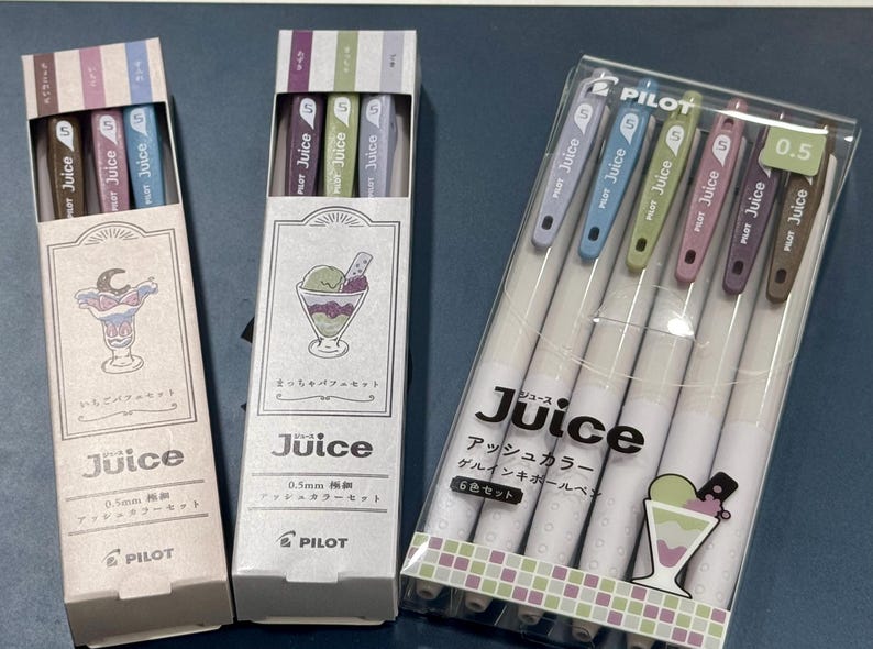 May include: Three sets of Pilot Juice 0.5mm gel ink pens in ash colors. Two sets are in vertical cardboard boxes with dessert illustrations, and one set is in a clear plastic case containing six pens with colored clips and white barrels.