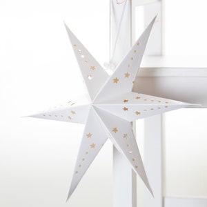 Pode incluir: Uma lanterna de estrela de papel branco com um padrão de estrelas douradas pendurada em uma cadeira branca. A estrela é dobrada e tem uma forma tridimensional.