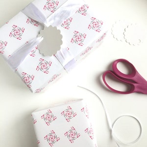 May include: Two white gift boxes wrapped in pink and gray patterned paper with white ribbon and a white tag. The boxes are on a white surface with a pair of pink scissors and two white scalloped circles.