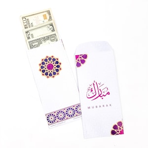 May include: Two white envelopes with pink and gold designs. One envelope features a geometric pattern and the other has the Arabic word "Mubarak" written in pink with the word "Mubarak" written in English below it. The envelope with the geometric pattern contains cash.