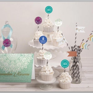 May include: A set of cupcakes decorated with Ramadan Kareem themed toppers. The cupcakes are white with white frosting and are arranged on a tiered cake stand. There is a card with the text "Ramadan Kareem" on the table. The cupcakes are decorated with various designs, including a lantern, a star, and a crescent moon.