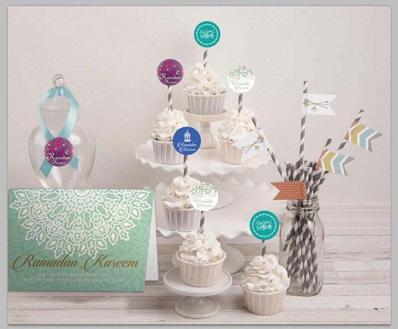 May include: A set of cupcakes decorated with Ramadan Kareem themed toppers. The cupcakes are white with white frosting and are arranged on a tiered cake stand. There is a card with the text "Ramadan Kareem" on the table. The cupcakes are decorated with various designs, including a lantern, a star, and a crescent moon.