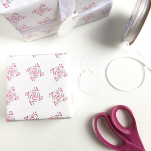 May include: A white gift box with a pink and gray patterned design, wrapped with a white ribbon and a white gift tag. The box is sitting on a white surface next to a spool of white ribbon, a pair of pink scissors, and a white paper doily.