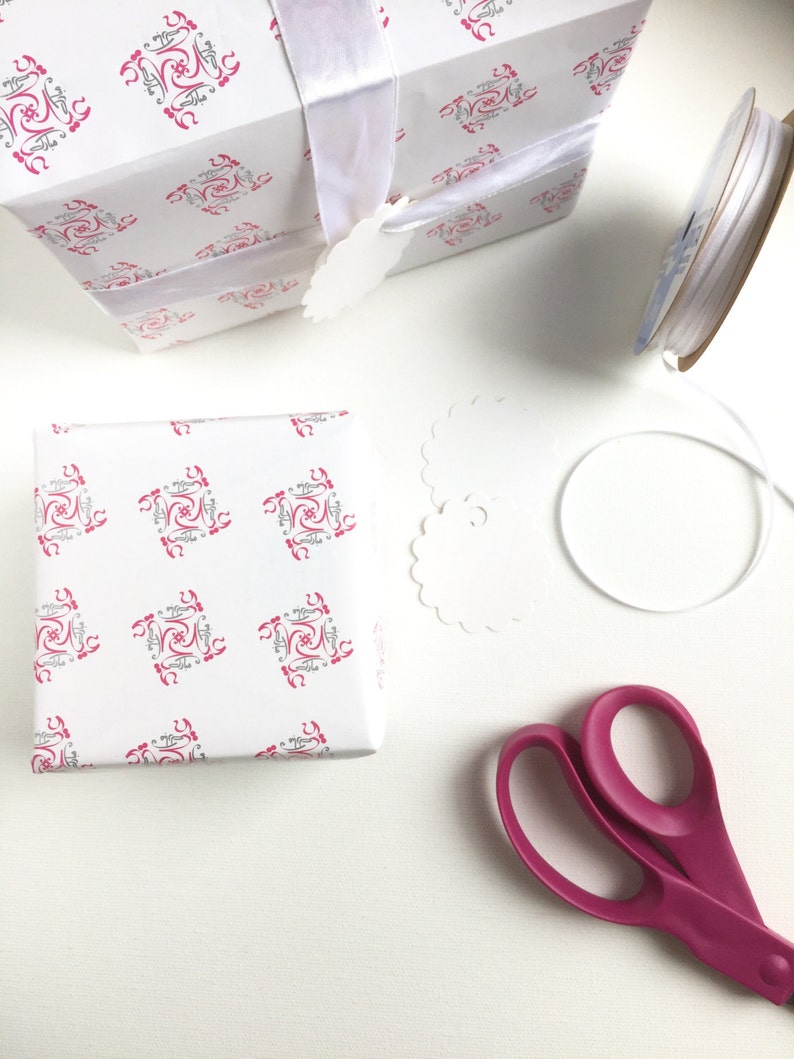 May include: A white gift box with a pink and gray patterned design, wrapped with a white ribbon and a white gift tag. The box is sitting on a white surface next to a spool of white ribbon, a pair of pink scissors, and a white paper doily.