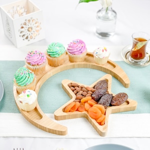 May include: A wooden crescent moon and star shaped serving tray with a variety of snacks. The tray features a crescent moon shape with a star in the center. The tray is filled with colorful cupcakes, almonds, dried apricots, and dates.