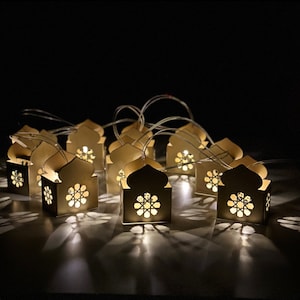 Może przedstawiać: Girlanda z 10 ozdobnych papierowych lampionów z białymi diodami LED. Każdy lampion ma kształt meczetu z misternymi wycięciami i złotym wykończeniem.