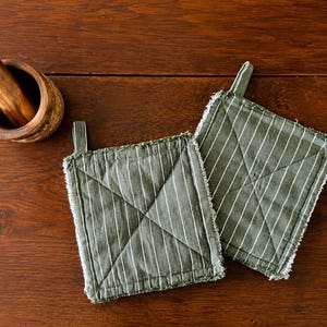 May include: Two square olive green pot holders featuring thin white vertical pinstripes, a quilted X-pattern across the center, and frayed edges with a small fabric hanging loop attached to the top corner of each piece, alongside a wooden mortar.
