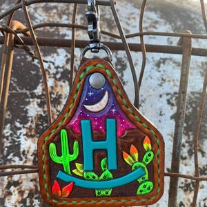 May include: A brown leather keychain with a painted desert scene. The design includes a teal letter "H", cacti, a crescent moon, and stars. The keychain has a silver-colored clasp.
