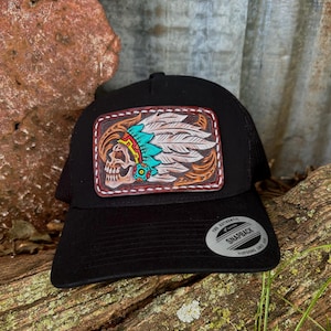 May include: Black trucker hat with a leather patch featuring a skull wearing a Native American headdress. The patch has turquoise, red, and white accents. The hat has a snapback closure and a mesh back.