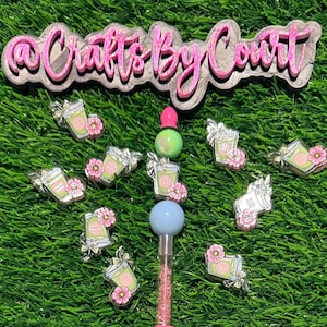 May include: A collection of decorative pins and a pen on a green surface. The pins feature a green drink with a pink flower and bow. The pen has a pink and clear barrel with a blue and green top. The text "Crafts By Court" is at the top.