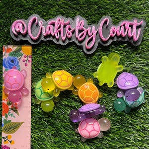 May include: A collection of colorful, translucent turtle-shaped charms in various colors, including pink, green, yellow, orange, and purple. The charms are arranged on a green surface, with a floral ruler and a pink and black sign that reads "@Crafts By Court".