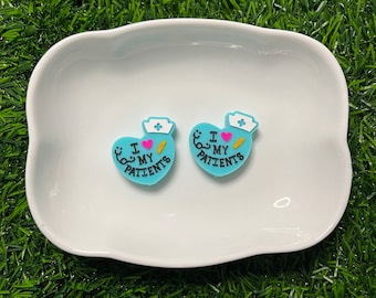 May include: Two blue heart-shaped charms with the text "I MY PATIENTS" in black, a nurse's hat, a stethoscope, and a syringe. The charms are on a white, wavy-edged dish, set against a backdrop of green artificial grass.