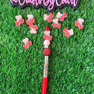 May include: A red and clear pen with a cherry-themed design, surrounded by cherry-shaped embellishments. The pen has a red barrel, a clear tube filled with colorful accents, and a decorative top. The text "@Crafts By Court" is visible.