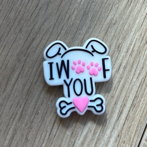May include: A white charm with black text that reads "I WOOF YOU". The charm features pink paw prints, a pink heart, and a bone design. The charm has a dog-like appearance with ears.