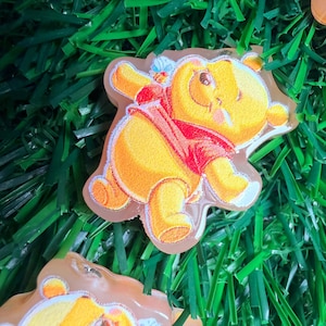 May include: A yellow Winnie the Pooh character patch with a red shirt and a light brown border. The patch is on a bed of green artificial grass. The character is in a walking pose.